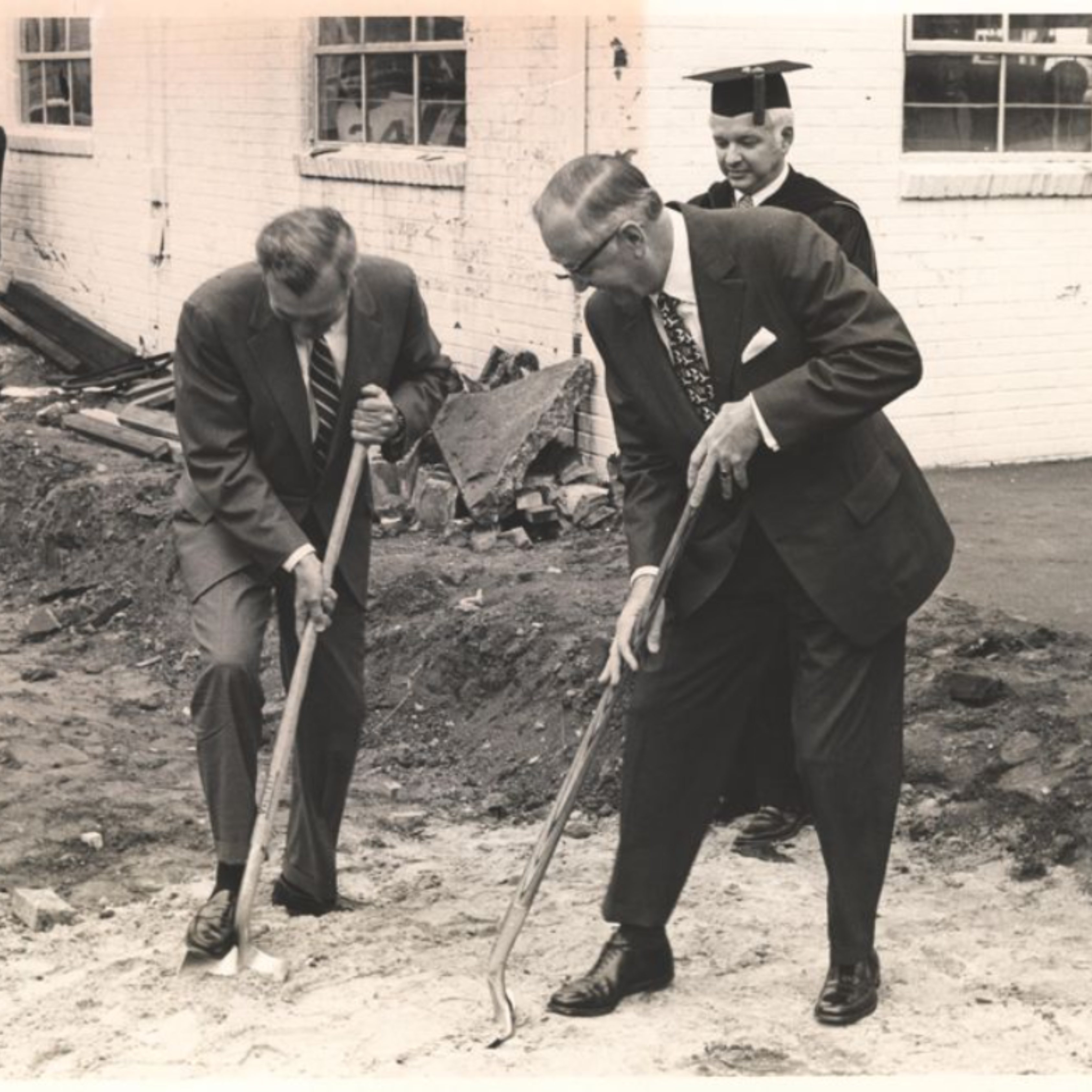 Two men in suits digging shovels into the ground, and a man standing behind them wearing academic regalia